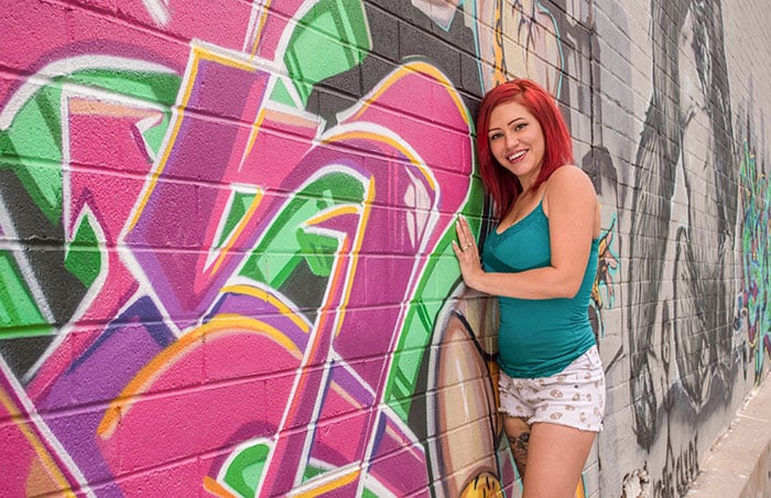 A photo of a redhaired woman smiling and leaning against a wall with street art on it.